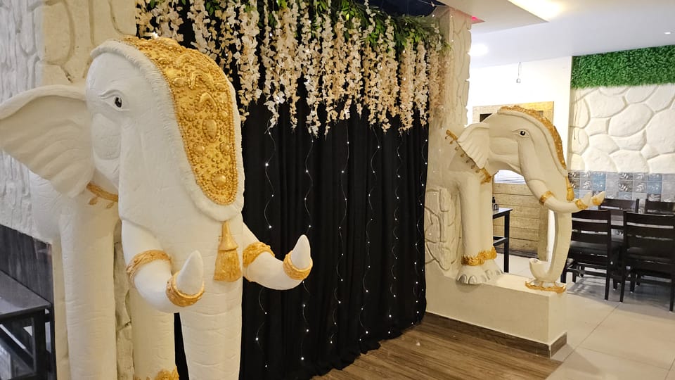 A white elephant statue draped in gold beside the entrance, with dining tables and a green foliage wall behind